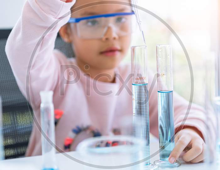 Asian Little Girl Holding And Dropping Blue Solution In Test Tube In Laboratory Room At Nursery Background. Science And Education Concept. Child And Kids Theme.