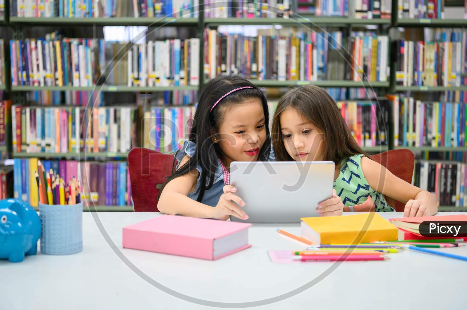 Two Little Happy Cute Girls Playing On A Tablet Pc Computing Device In Library At School. Education And Self Learning Wireless Technology Concept. People Lifestyles And Friendship. Preschool Children