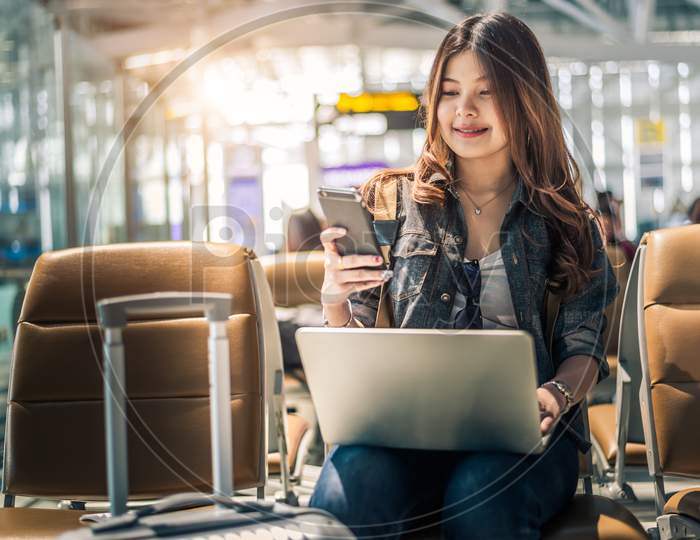 Young Asian Female Passenger Using Laptop And Smart Phone While Sitting On Seat In Terminal Hall And Waiting For Flight In Airport . People Lifestyles And Happy Woman. Technology And Travel Concept.
