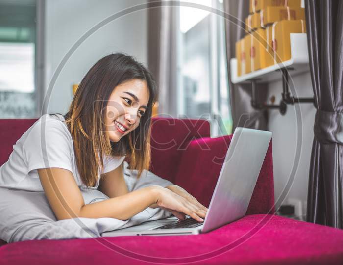 Young Woman Surfing The Internet By Laptop On Red Sofa In Cheerful Gesture Mood Emotion. Selling And Online Shopping Concept. Merchant And Marketing. Happiness Of New Business Trader  And Entrepreneur