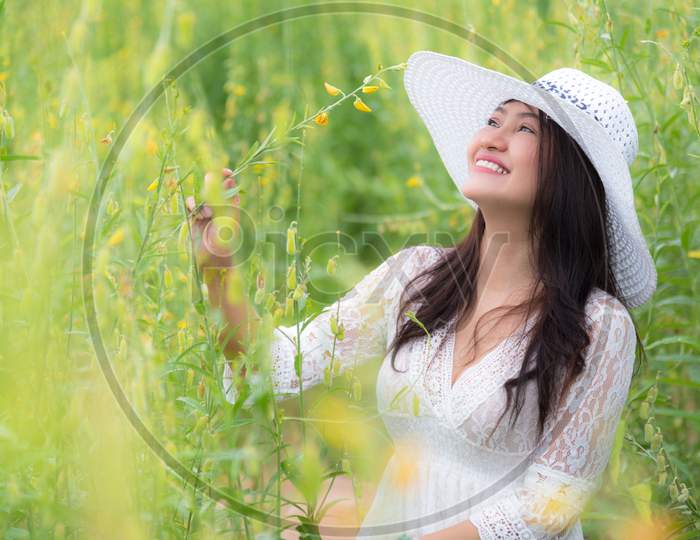 Beauty Asian Woman In White Dress And Wing Hat Walking In Rapeseed Flower Field Background. Relaxation And Travel Concept. Wellness Happy Life Of Girl On Vacation. Nature And People Lifestyle