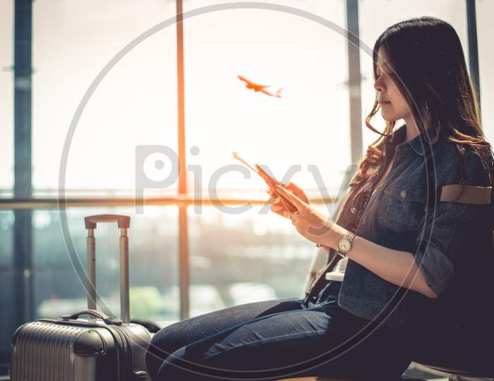 Beauty Asian Woman With Suitcase Luggage Waiting For Departure While Using Smart Phone In Airport Lounge. Female Traveler And Tourist Theme. High Season And Vacation Concept. Airplane Background