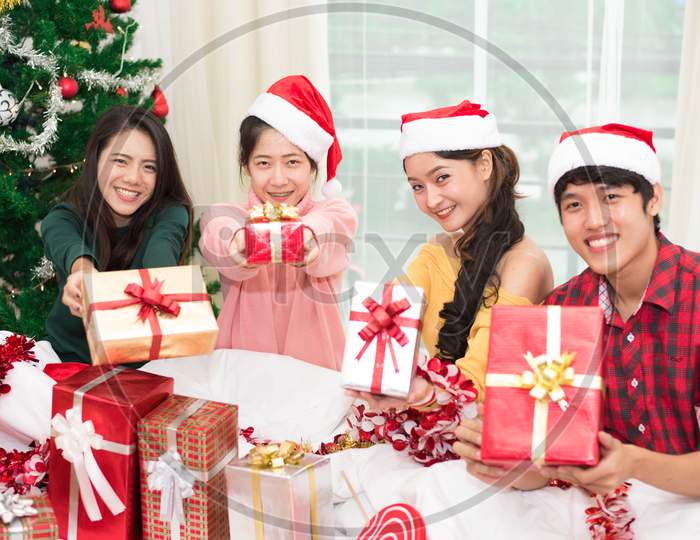 Group Of Young Asian People Holding And Giving Gift Box To You With Xmas Tree Background. Holiday And Festival Concept. Christmas And New Year Event Theme. Happy People Wearing Santa Hat.