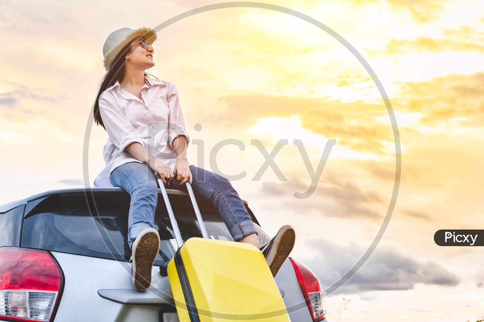 Happy Asian Woman On Top Of Car With Luggage Bag. Girl Sitting On Roof And Looking Sunset Before Night In Evening. People Lifestyle In Long Vacation Trip Concept. Nature And Transportation Vehicle.