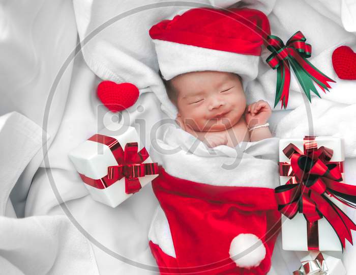 Sleeping Newborn Baby Face In Christmas Hat With Gift Box From Santa Claus And Yarn Heart On White Soft Towel. Cute Infant Lifestyle And Innocent Happy Baby Lying In Cold Snow Season. New Year Winter