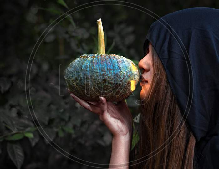 Female Wizard Kissing Pumpkin Lantern. Female Costume In Black Clothes And Hood On Mystery Forest Background. Halloween Day And Mystery Concept. Fantasy Of Magic Theme. Afterlife And Death Concept.