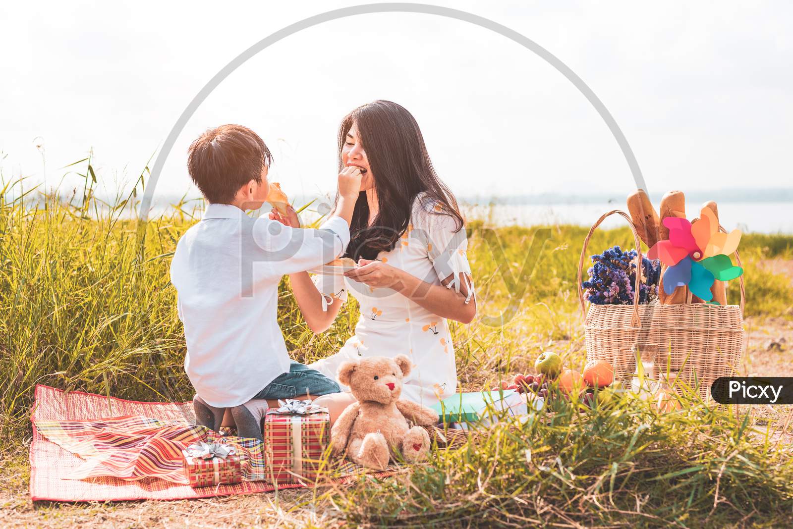 Little Asian Boy His Mom Feeding Snack Each Other In Meadow When Doing Picnic. Mother And Son Playing Together. Celebrating In Mother Day And Appreciating Concept. Summer People And Lifestyle Theme.