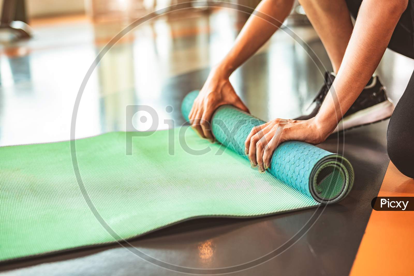 Close Up Of Sporty Woman Folding Yoga Mattress In Sport Fitness Gym Training Center Background. Exercise Mat Rolling Keeping After Yoga Class. Workout And Sport Training Concept. Hands On Carpet