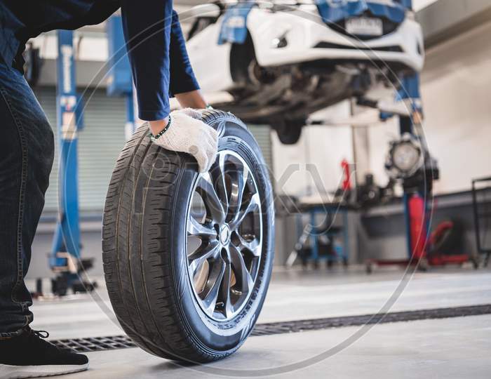 Male Mechanic Hold And Rolling Tire At Repairing Service Garage Background. Technician Man Replacing Winter And Summer Tyre For Safety Road Trip. Transportation And Automotive Maintenance Concept