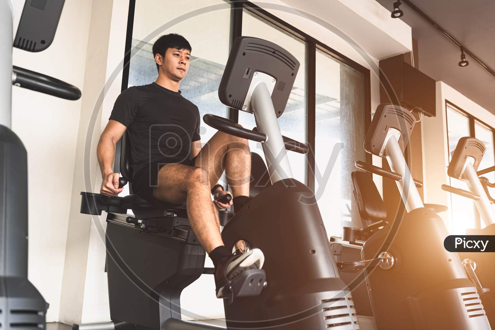 Asian Young Sport Man Riding Stationary Bicycle In Fitness Gym. Man Working Out On Spinning Bikes In Gym. People Lifestyles And Health Club Concept. Bodybuilding Theme