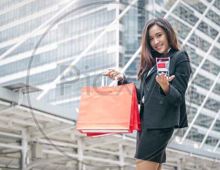 Beauty Asian Woman Holding Shopping Bag And Shopping Cart In Mall.  Shopaholic In Black Friday And Cyber Monday Sale Concept. Businesswoman Posing Product Present In Urban Or Metropolis As Shop Buyer.