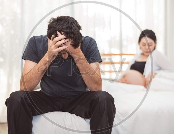 Worried Stress Man Sitting On Bed With Hand On Forehead In Bedroom In Serious Mood Emotion With Pregnant Wife Woman Background. Major Depressive Disorder Called Mdd Concept. Physical Healthcare