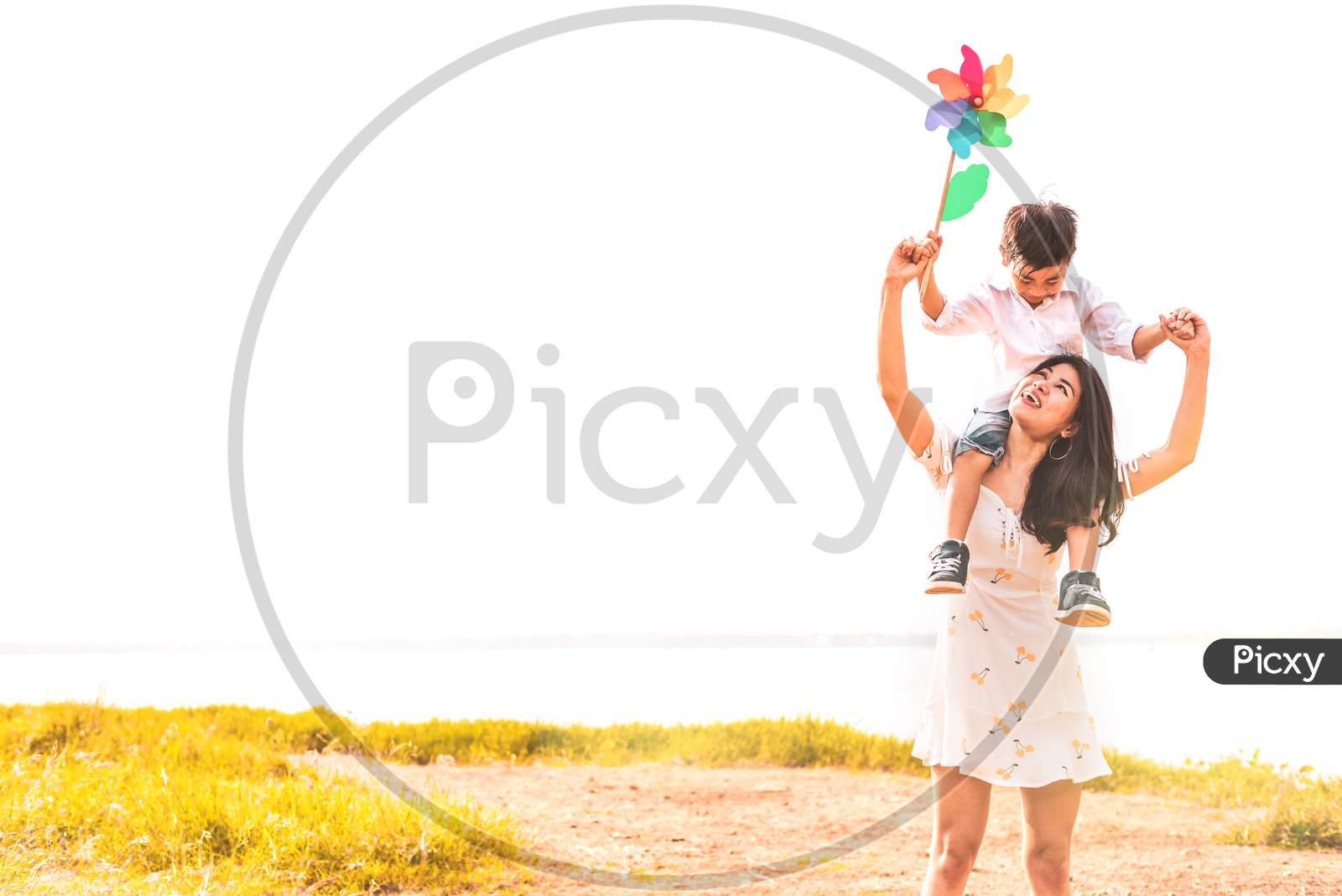 Little Asian Boy Riding Back On His Super Power Mom In Meadow Near Lake. Mother And Son Playing Together. Celebrating In Mother Day And Appreciating Concept. Summer People And Lifestyle Theme.