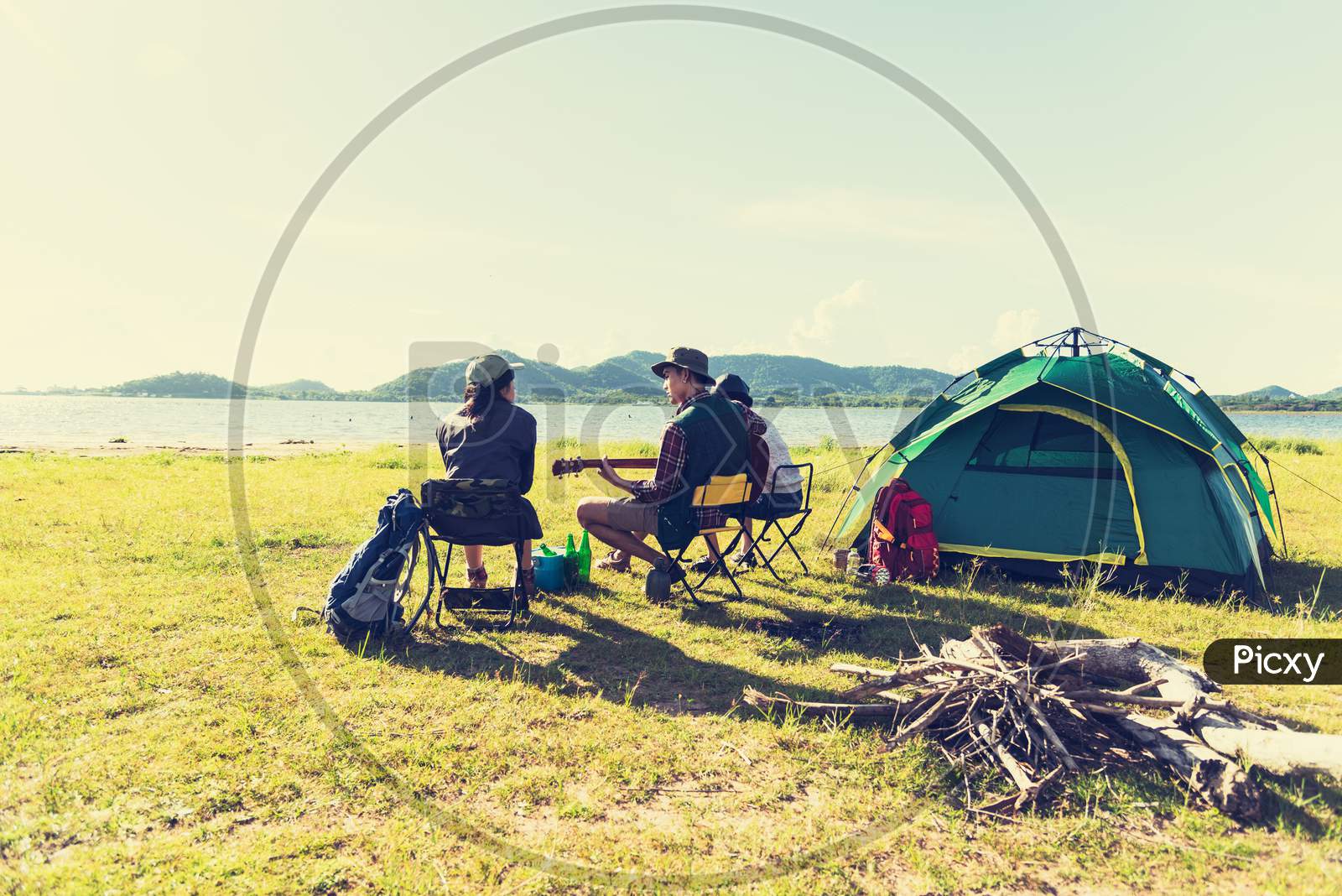 Group Of Travelers Camping And Doing Picnic And Playing Music Together. Mountain And Lake Background. People And Lifestyle. Outdoors Activity And Leisure Theme. Backpacker And Hiker. Back View Angle
