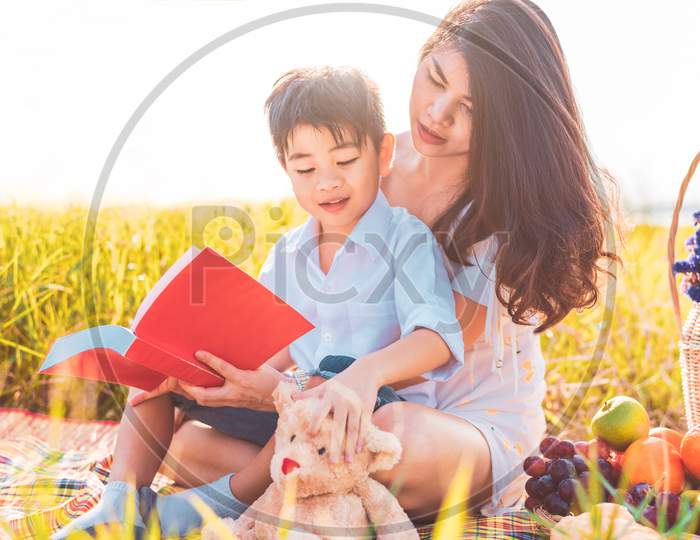 Little Asian Boy And His Mother Reading Books When Doing Picnic In Meadow. Mother And Son Learning Together. Celebrating In Mother Day And Appreciating Concept. Summer People And Lifestyle Education