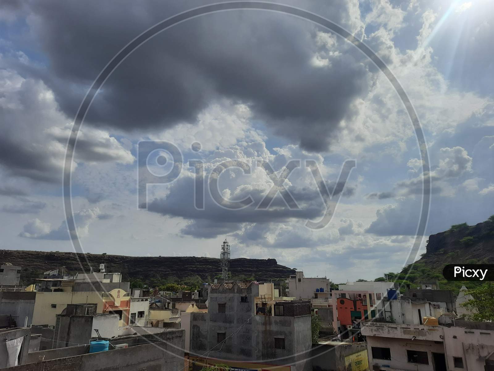 Dark and white clouds, blue sky behind, sun Ray's travelling through the clouds, small mountain and buildings at the surface, beautiful natural scene