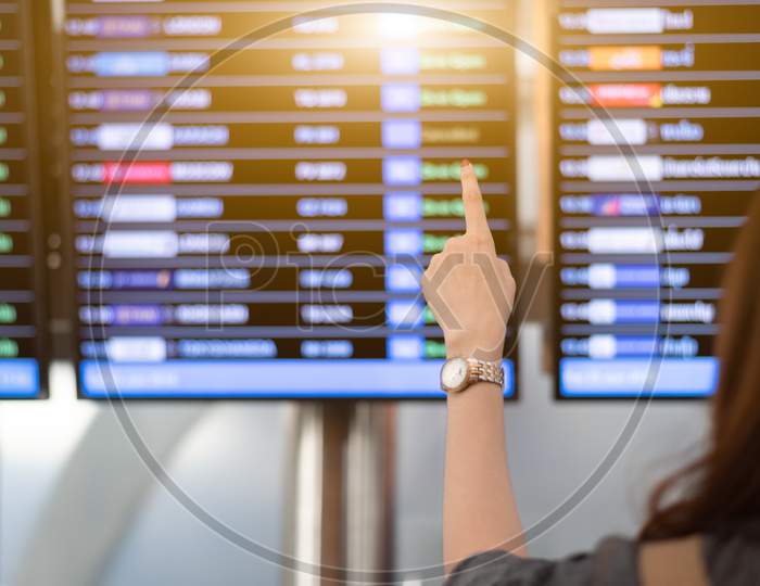 Back View Of Woman Looking For Flights From Flight Schedule In Airport. Female Tourist Pointing At Time Table For Take Off Plane. Travel And Transportation Concept. Vacation And Long Holiday Theme.