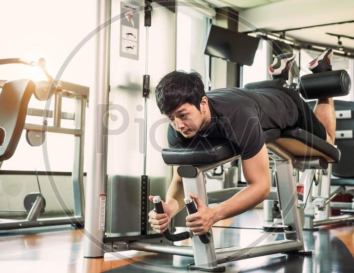 Asian Sport Man Stretching And Lifting Weight By Two Legs When Facing Down For Stretching Muscle At Fitness Gym At Condominium Background. Sport And People Lifestyles Concept. Leg Curl Posture