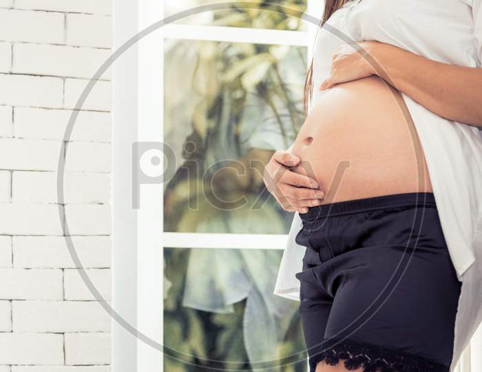 Close Up Belly Of Pregnant Woman In Home Interior And Window Background. Healthy And Maternity Parent Concept. Mother Take Care Of Her Child In Belly. Happy Life Of Mom Before Maternity