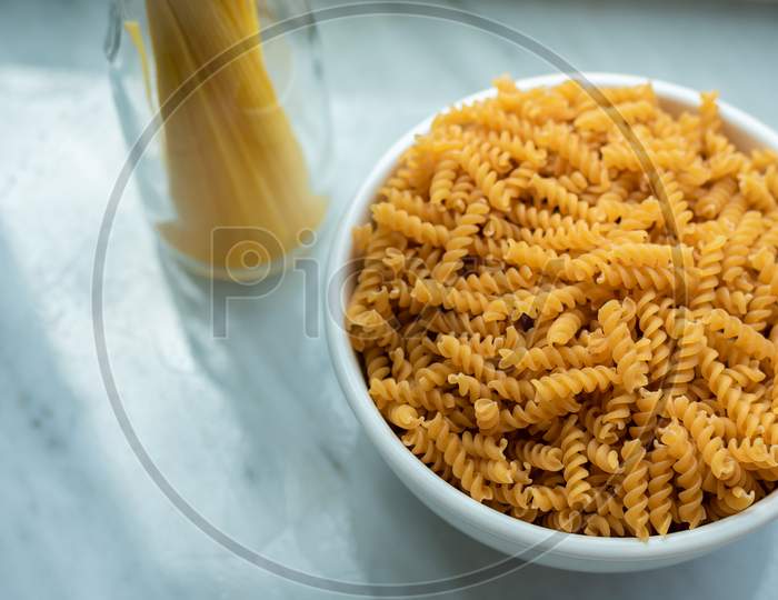Top View Of White Bowl Of Spiral Macaroni And Tied Spaghetti Stand Erect At Windows Of Kitchen Room. Food And Ingredients Concept. Italian Food Theme