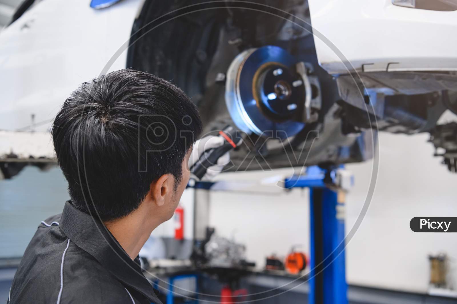 Asian Male Mechanical Hold And Shining Flashlight To Examine Car Disk Brake Pad Wear Of Automotive Vehicle. Safety Suspension Inspection Check Service Maintenance For Customer Before Road Trip Concept