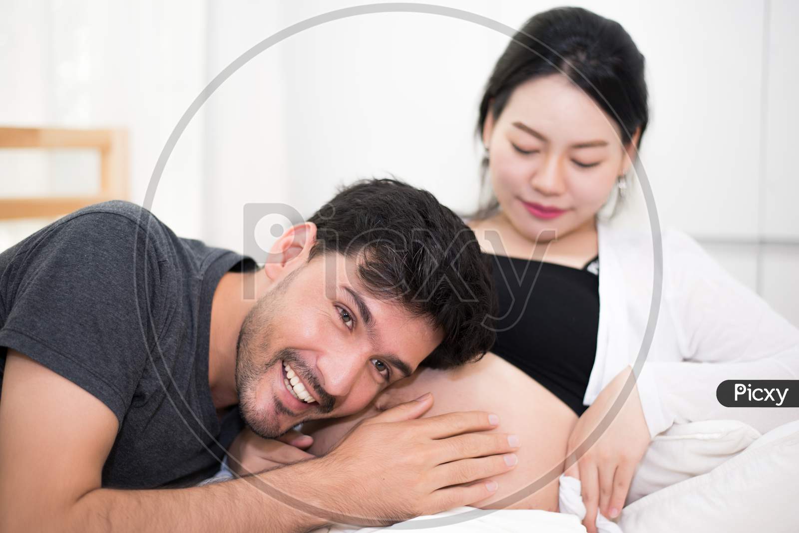 Father Hearing His Son Or Daughter Kicking Sound Inside Mother Belly When Sitting On Lying On Bed At Home. Family And Lovers Concept. Happy Sweet Home And Wedding Theme