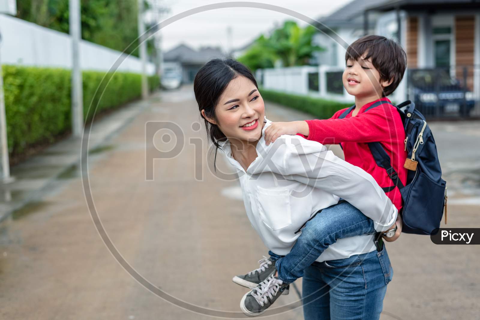 Single Mom Carrying And Playing With Her Children Near Home With Villa Street Background. People And Lifestyles Concept. Happy Family And Home Sweet Home Theme. Outdoors And Nature Theme.
