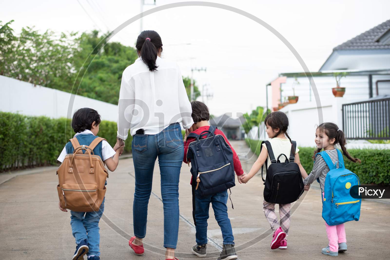 Group Of Preschool Student And Teacher Holding Hands And Walking To Home. Mom Bring Her Children Go To School Together. Back To School And Education Concept. People And Lifestyles Theme. Back View