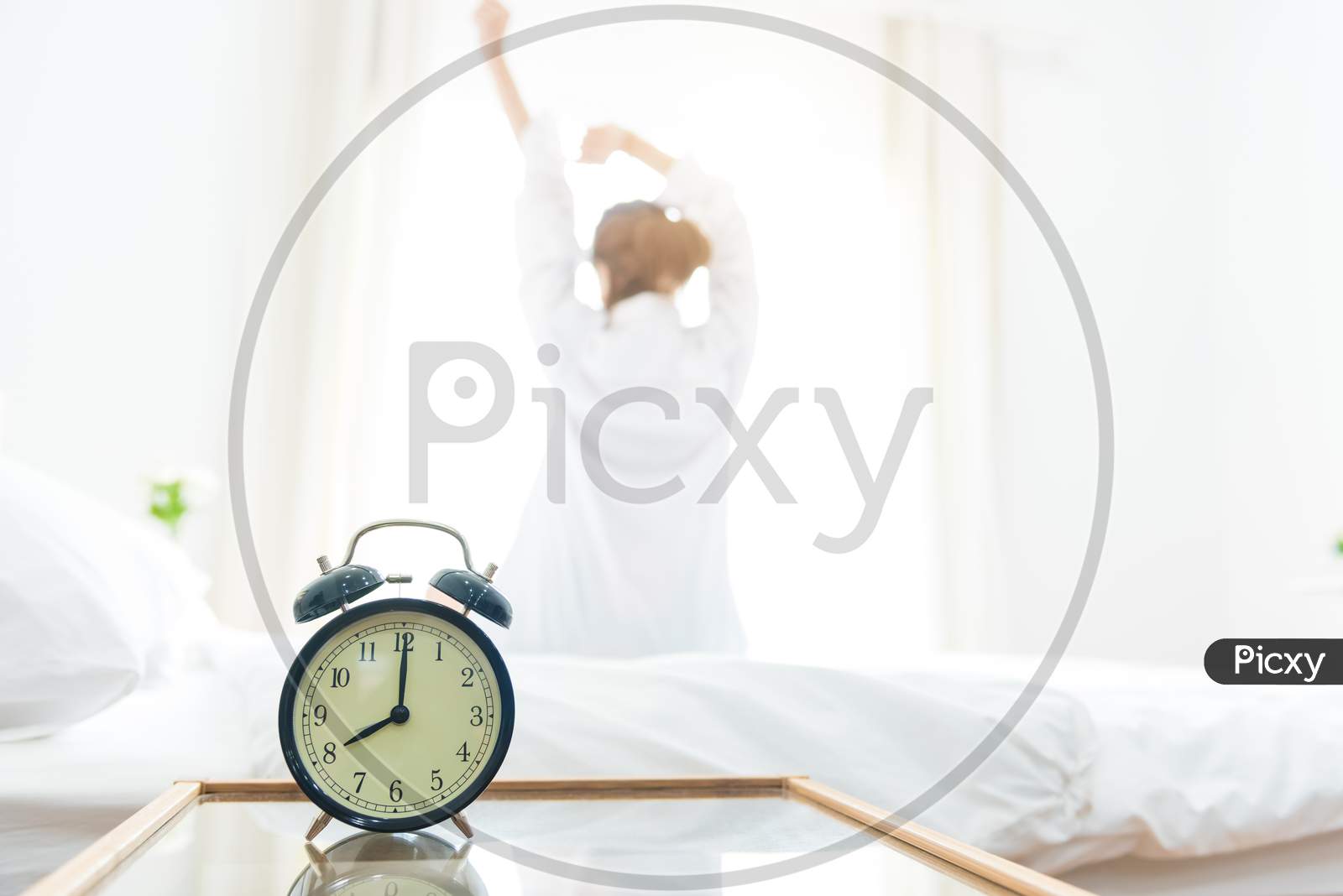 Back View Of Woman Stretching In Morning After Waking Up On Bed Near Window With Alarm Clock. Holiday And Relax Concept. Lazy Day And Working Day Concept. Office Woman And Worker In Daily Life Theme