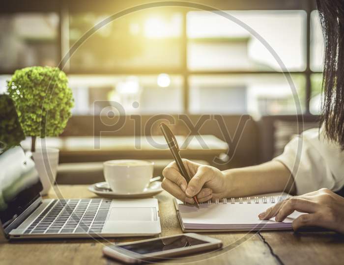 Close Up Of Woman Hand Writing Business Project Planning In Notebook With Laptop And Smart Phone At Coffee Shop. People And Technology Concept. Freelance And Lifestyle Theme. Entrepreneur And Working