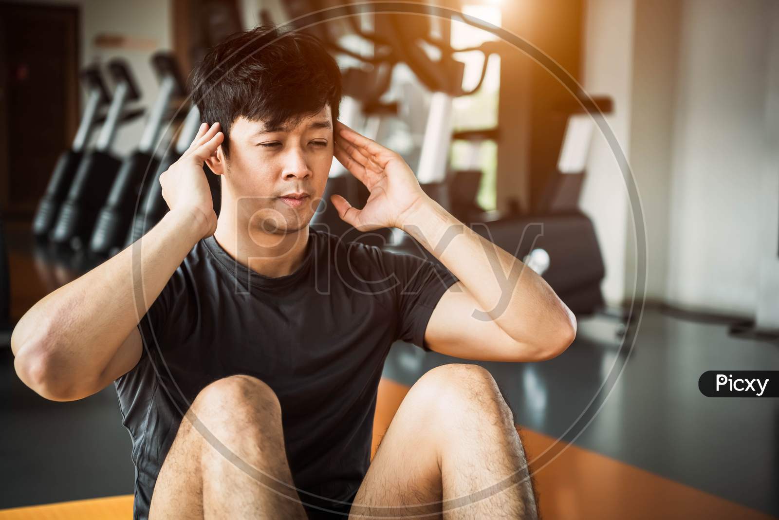 Asian Sport Man Doing Crunch Or Sit Up Posture On Yoga Mat In Fitness Gym At Condominium With Gym Equipment Background. Office Working People Lifestyles And Sport Workout Concept.