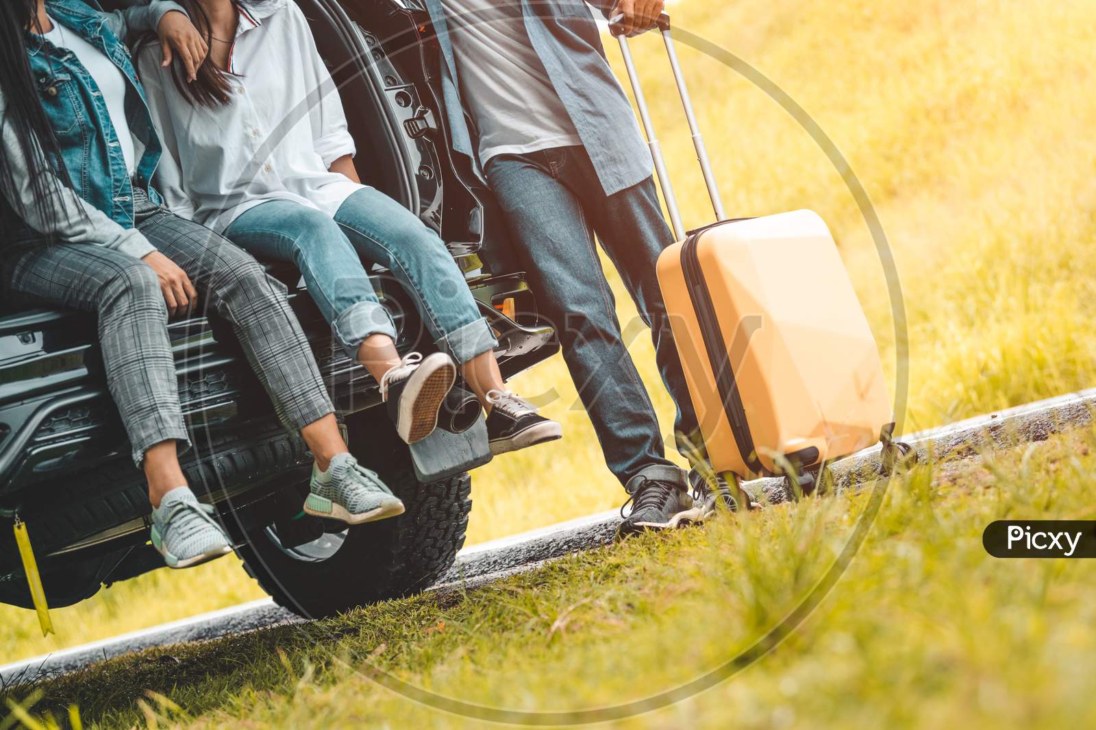Closeup Lower Body Of Group Of Friends Relaxing On Suv Car Trunk With Trolley Luggage Along Road Trip With Autumn Mountain Hill Background. Freedom  Road Way. People Lifestyle Transportation Travel