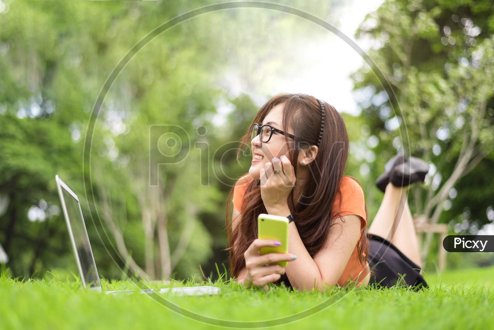 Happy Asian Woman Resting And Looking Beside In Park With Smartphone. People And Lifestyles Concept. Technology And Beauty Theme.
