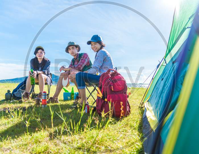 Group Of Travelers Camping And Doing Picnic In Meadow With Tent Foreground. Mountain And Lake Background. People And Lifestyles Concept. Outdoors Activity And Leisure Theme. Backpacker And Hiker Theme