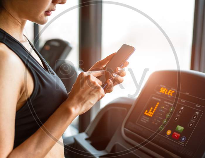 Woman Using Smart Phone When Workout Or Strength Training At Fitness Gym On Treadmill. Relax And Technology Concept. Sports Exercise And Health Care Theme. Happy People And Comfortable Application