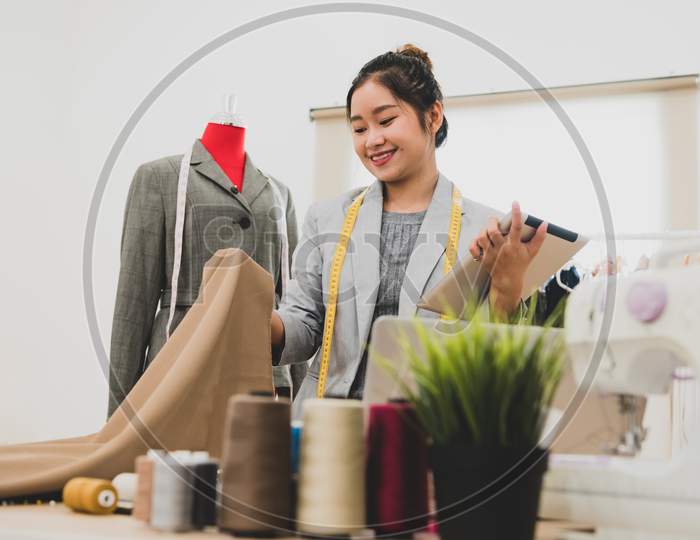 Fashion Designer Stylist In Business Owner Workshop With Tablet And Customer Contact List. Tailor And Sew Concept. Portrait Of Happy Casual Trendy Fashion Businesswoman In Studio. Job And Occupation