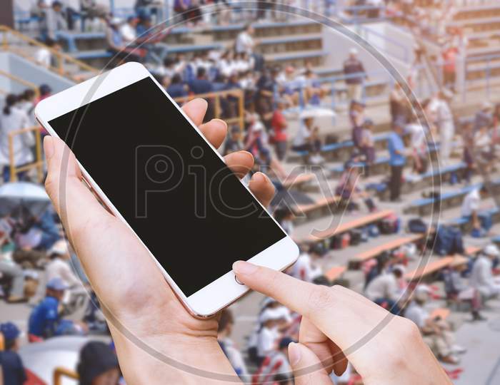 Woman Hold Smart Phone And Touch Button By Hand With Blank Screen For Advertisement, Stadium And Audiences Background ,Technology And Sport Concept