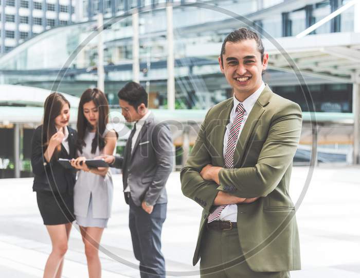 Smiling Young Businessman Looking Into Camera With His Arms Crossed And Teammates Background In Urban. Modern Business Office Team At Outdoors In City. Business People And Leadership Lifestyle Concept