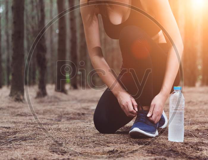 Woman Tying Up Shoelaces When Jogging In Forest Back With Drinking Water Bottle Beside Hers. Sneakers Rope Tying. People And Lifestyles Concept. Healthcare And Wellness Theme. Park And Outdoors Theme.