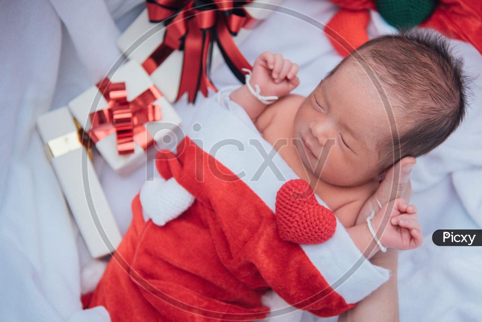 Sleeping Newborn Baby On Mother Hand In Christmas Hat With Gift Box From Santa Claus And Yarn Heart On White Soft Towel. Cute Infant Lifestyle And Innocent Lying In Cold Snow Season. New Year Winter