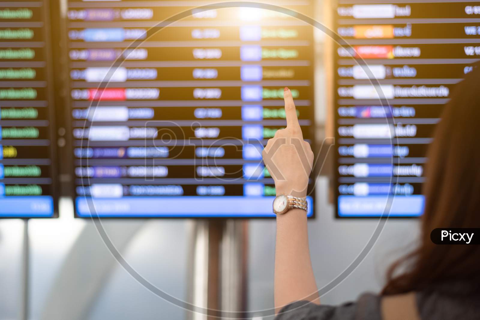 Back View Of Woman Looking For Flights From Flight Schedule In Airport. Female Tourist Pointing At Time Table For Take Off Plane. Travel And Transportation Concept. Vacation And Long Holiday Theme.