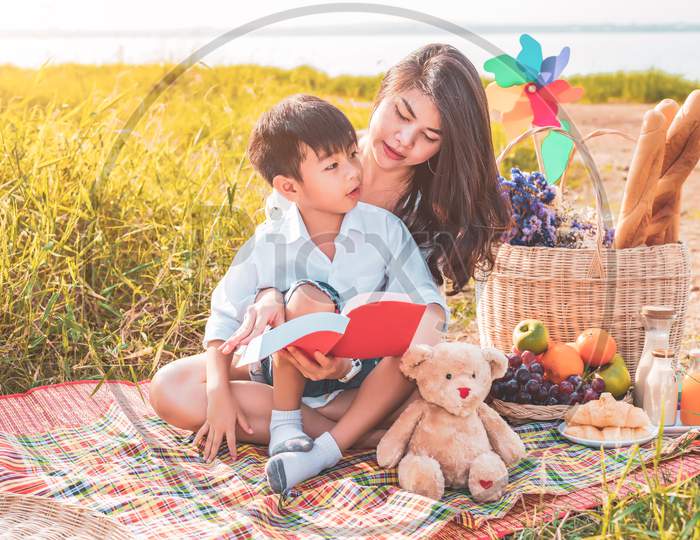 Beautiful Asian Mother And Son Doing Picnic And Reading Tale In Red Books In Easter Summer Party On Meadow Near Lake And Mountain. Holiday Vacation. People Lifestyle And Happy Family Life. Thai Person