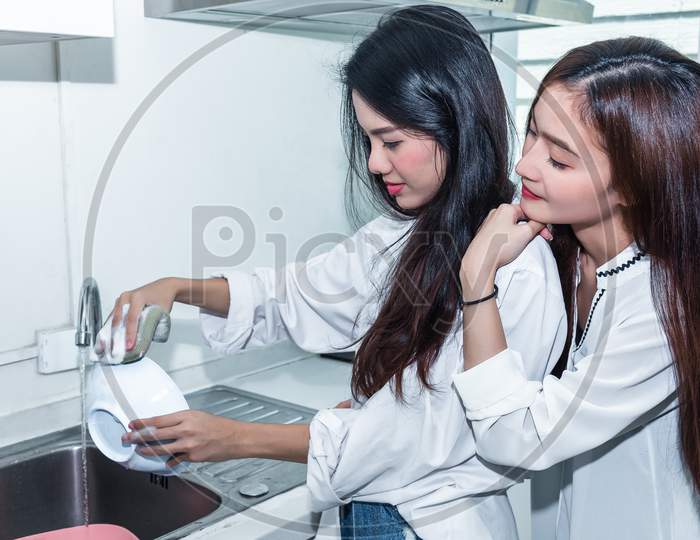 Two Asian Women Washing Dishes Together In Kitchen. People And Lifestyles Concept. Lgbt Pride And Lesbians Theme.