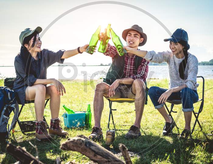 Group Of Young Asian Friends Enjoy Picnic And Party At Lake With Camping Backpack And Chair. Young People Toasting And Cheering Bottles Of Beer. People And Lifestyles Concept. Outdoor Background Theme