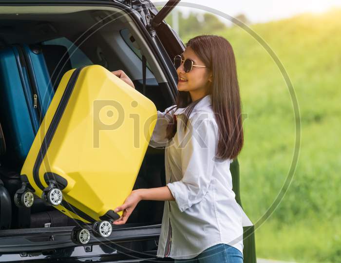 Asian Woman Lifting Yellow Suitcase Into Suv Car During Travel In Long Weekend Trip. People Lifestyles And Transportation Concept. Girl Put Luggage From Car Trunk To Camping. Selective Focus On Car