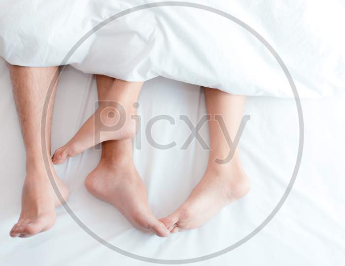 Closeup Feet Of Couple On The Bed. Man And Woman Lovers Make Love Under The Blanket Or Bed Sheet. Sex On Vacation Theme. Valentine And Honeymoon Concept.