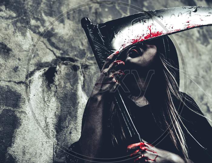 Witch Licking Blood On Reaper. Female Demon Angel In Black Clothes And Hood On Grunge Wall Background. Halloween Day And Mystery Concept. Fantasy Of Magic Theme. Afterlife And Death Concept.