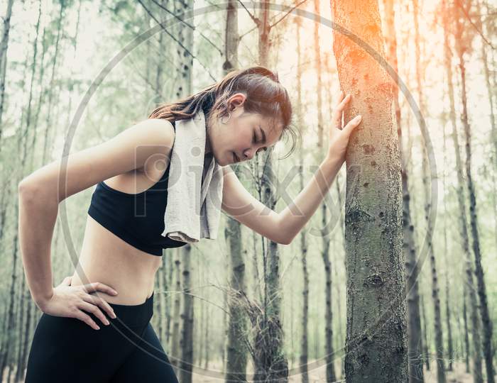 Asian Beauty Woman Tiring From Jogging In Forest. Lean On Tree. Towel And Sweat Elements. Sport And Healthy Concept. Jogging And Running Concept. Relax And Waist Pain Theme. Outdoors Activity Theme.