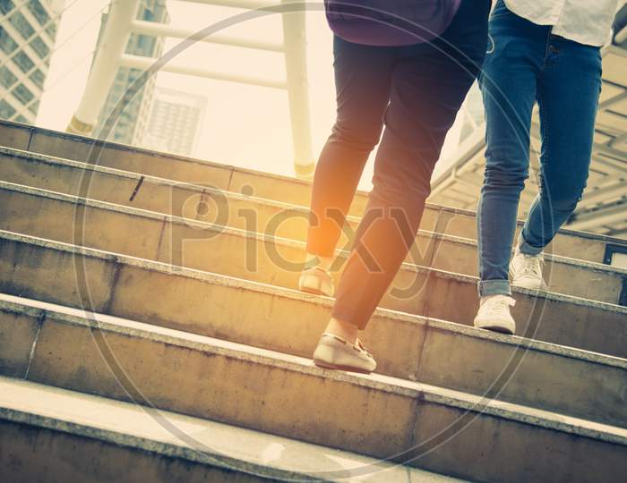 Close Up Legs Of Two Traveling People Walking On Stepping Up Stair In Modern City. Sneakers And Jeans Elements. Business And Travel Concept. City Lifestyle And Working People Theme. Rush Hours Theme.