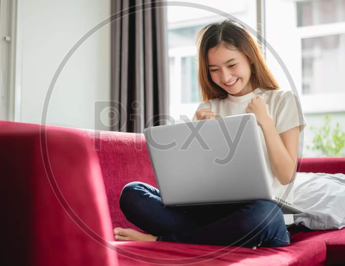 Young Woman Surfing The Internet By Laptop On Red Sofa And Glad In Successful Trading In Her House. Selling And Online Shopping Concept. Merchant And Marketing Concept. Happiness Of New Trader Theme